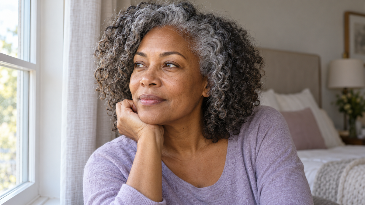 Mature Black woman with salt-and-pepper curls and natural eyebrows seated beside a bright bedroom window