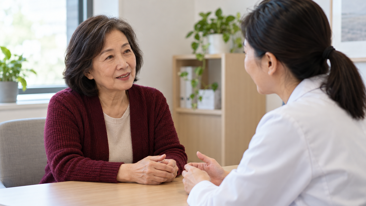 Mature East Asian woman in a burgundy cardigan speaking with a clinician across a consultation desk